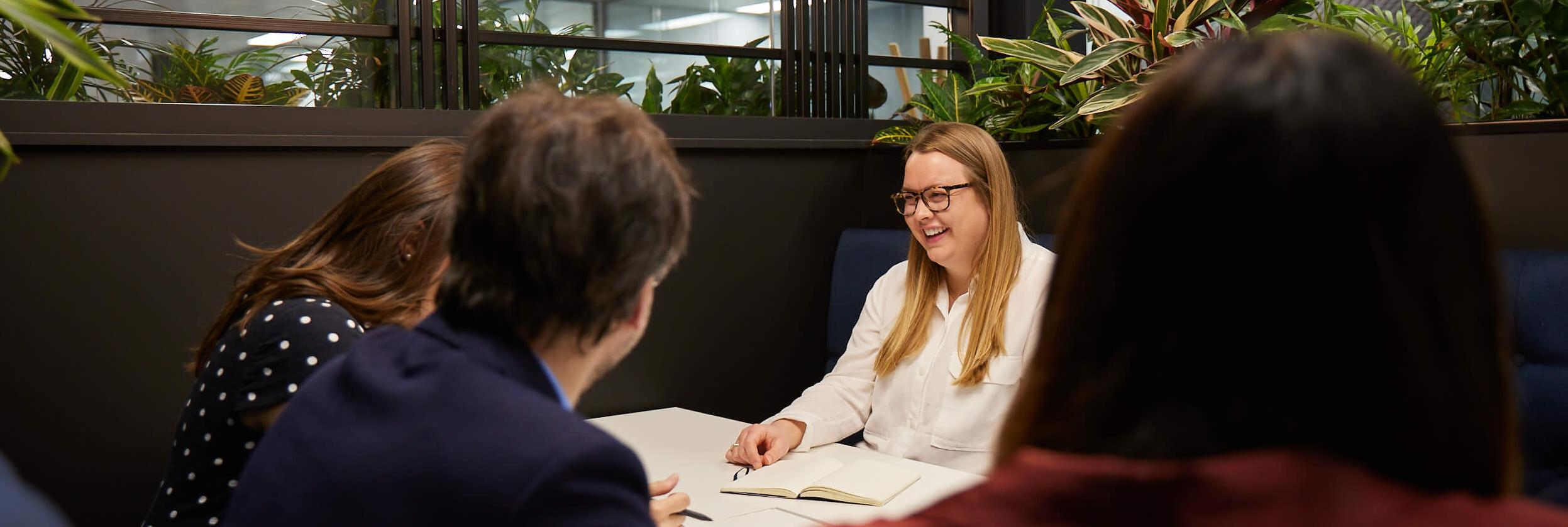 A woman in glasses smiles during a meeting with colleagues in a modern office.