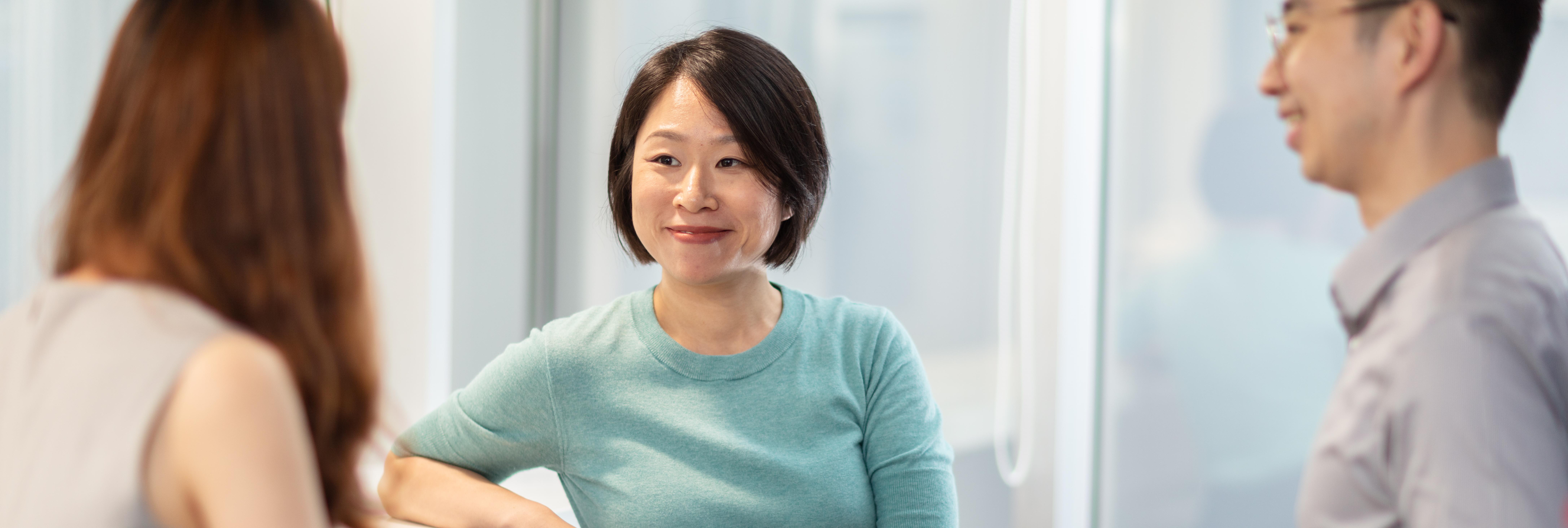 A woman with short dark hair in a teal top smiles and looks towards someone off-camera, with parts of two other individuals visible on either side of the frame.